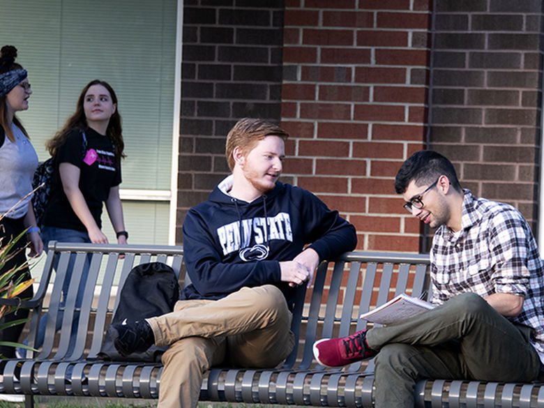 Two students studying on a bench, and students walking behind them. 