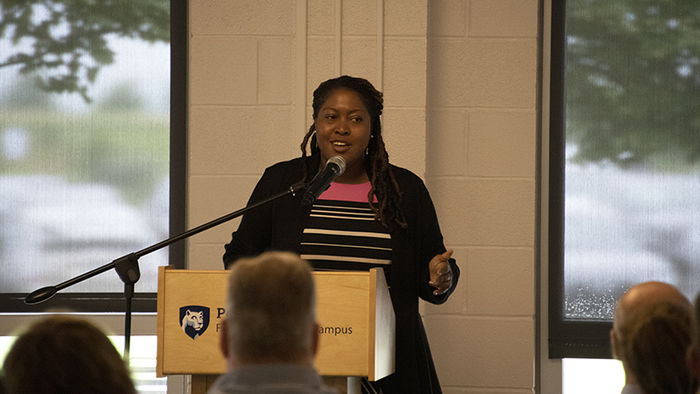 A woman in a black blazer speaks at a podium to a crowd of Penn State affiliates.