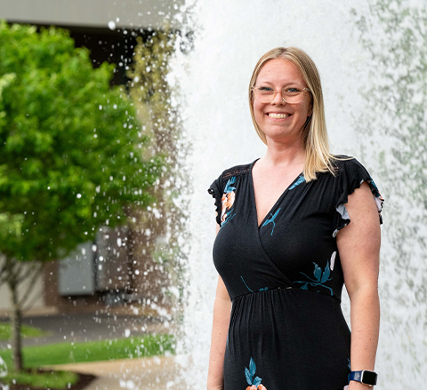 Amanda Collins stands smiling in front of a campus fountain, wearing a black floral dress and glasses.