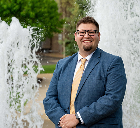 Barry Pawloski stands in front of a fountain on campus, wearing a blue suit, yellow tie, and glasses, smiling at the camera.