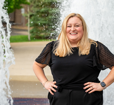 Holly Smallwood smiles broadly in front of a campus fountain, wearing a black outfit with sheer sleeves and a smartwatch.