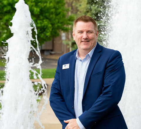 Jay Arcuri stands smiling beside a campus fountain, wearing a blue suit jacket and patterned shirt with a name badge.