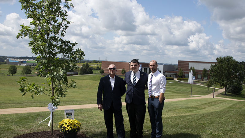 Three men stand on campus beside a commemorative maple tree to honor the memory of Trevor Thomas.