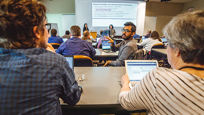 A group of people seated with laptops looking  at two presenters standing in the front of the room with an overhead presentation on the screen