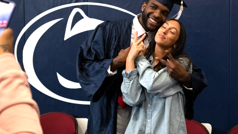 A graduate in cap and gown poses with a woman flashing peace signs as they smile for a photo in front of a Penn State backdrop.