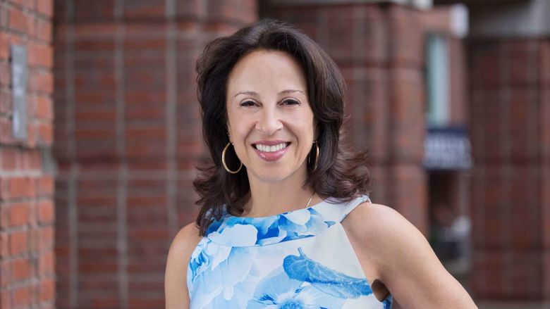 Maria Hinojosa wears a blue and white top and smiles at the camera in front of a brick structure