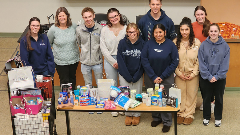 Group of Penn State Fayette students and staff standing behind a table and cart filled with donated hygiene items including toothpaste, shampoo, soap, and other personal care products.