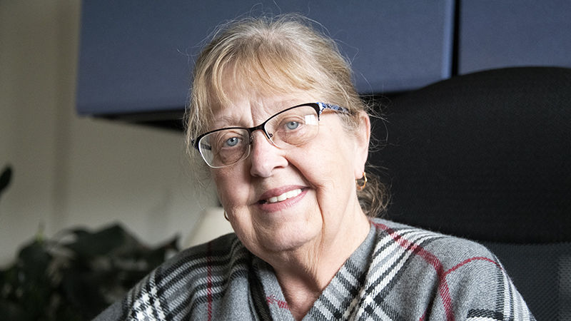 A woman, Mary Ann Walters, is seated at her desk at Penn State Fayette. She wears a striped shawl and glasses.