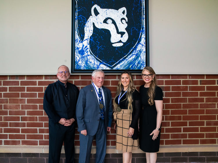 W. Charles Patrick, Charles Hunnell, Kelsey Beckas, and Tiffany Guittap posing in front of framed artwork of the Nittany Lion.
