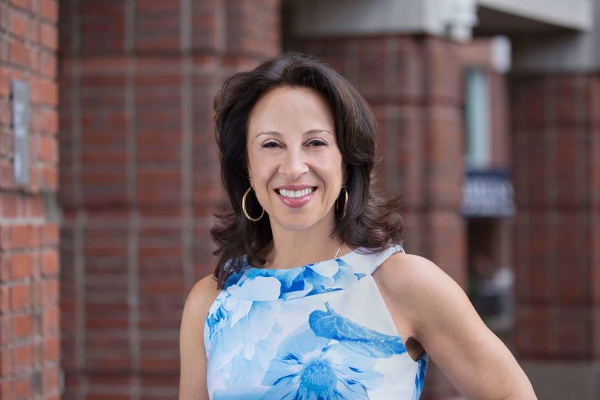 Maria Hinojosa wears a blue and white top and smiles at the camera in front of a brick structure